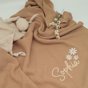 Puede incluir: Una manta de beb&eacute; de punto color canela con el nombre "Sophia" bordado en cursiva, junto con flores de margarita blancas. Un conejito de peluche y una mantita de seguridad de muselina est&aacute;n sobre la manta. Tambi&eacute;n hay un peque&ntilde;o ramo de flores secas.