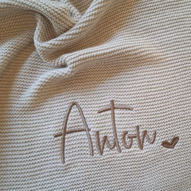 Puede incluir: Una manta beige tejida con el nombre "Anton" bordado en hilo marr&oacute;n a juego. Un peque&ntilde;o coraz&oacute;n est&aacute; bordado junto al nombre. La manta est&aacute; doblada y tiene un aspecto texturizado.