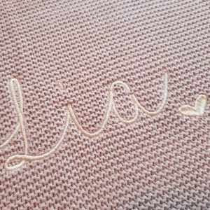 Puede incluir: Primer plano de una manta de punto en un tono rosa suave. La palabra "Lucy" est&aacute; bordada en hilo crema, con un peque&ntilde;o coraz&oacute;n al final. La textura del tejido de punto es visible.