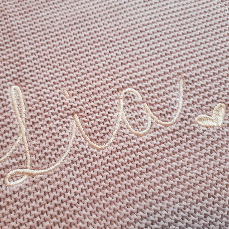 Puede incluir: Primer plano de una manta de punto en un tono rosa suave. La palabra "Lucy" est&aacute; bordada en hilo crema, con un peque&ntilde;o coraz&oacute;n al final. La textura del tejido de punto es visible.