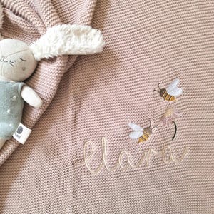 Könnte beinhalten: Eine altrosa Strickdecke mit einer Stickerei aus zwei Bienen und einer Blume sowie dem in Schreibschrift gestickten Namen "Elara". Ein Plüschhase mit langen, flauschigen weißen Ohren und einem salbeigrünen Outfit liegt auf der Decke.