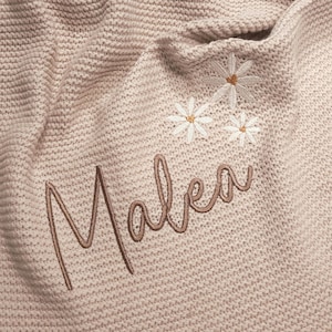Puede incluir: Una manta de punto beige claro con el nombre "Malea" bordado en cursiva. La manta presenta tres margaritas bordadas con p&eacute;talos blancos y centros amarillos. La manta est&aacute; hecha de un material suave y texturizado.