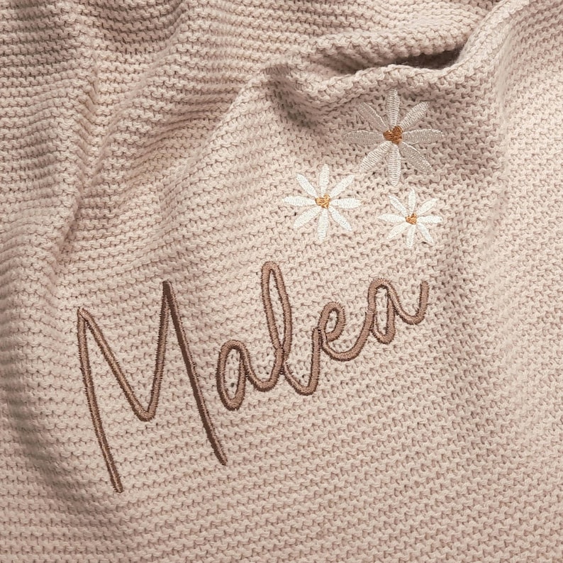Puede incluir: Una manta de punto beige claro con el nombre "Malea" bordado en cursiva. La manta presenta tres margaritas bordadas con p&eacute;talos blancos y centros amarillos. La manta est&aacute; hecha de un material suave y texturizado.