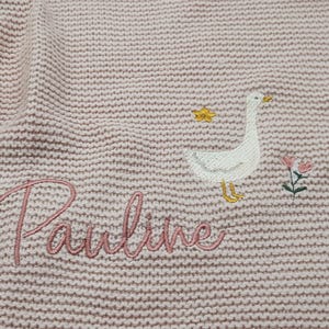 Op de afbeelding: Een lichtroze gebreide stof met een geborduurde witte gans met een gele snavel en poten, een kleine gele ster, een roze en groene bloem, en de naam Pauline geborduurd in een cursief roze lettertype.