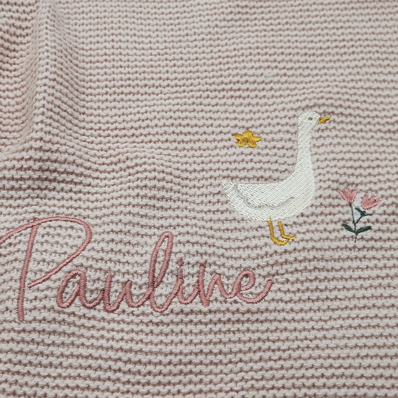 Op de afbeelding: Een lichtroze gebreide stof met een geborduurde witte gans met een gele snavel en poten, een kleine gele ster, een roze en groene bloem, en de naam Pauline geborduurd in een cursief roze lettertype.