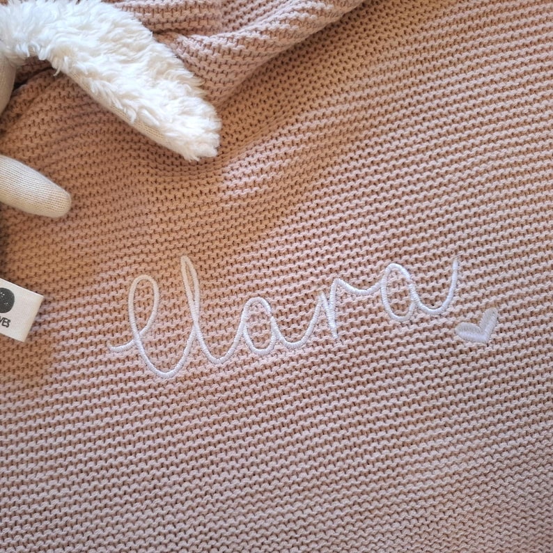 Puede incluir: Una manta de punto rosa claro con el nombre "Lara" bordado en escritura blanca. Se ve una oreja de conejo de peluche blanco y parte de la cabeza del conejo. La manta tiene un aspecto texturizado.