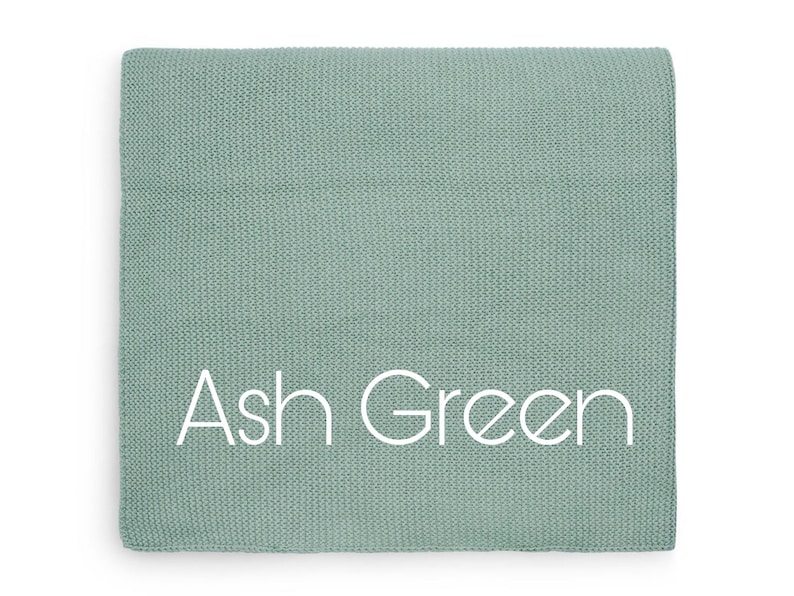 Puede incluir: Una manta cuadrada y doblada en color verde ceniza apagado. La manta tiene un patr&oacute;n de tejido texturizado. Las palabras "Ash Green" est&aacute;n impresas en blanco en la parte delantera de la manta.