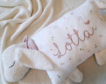 Puede incluir: Un juguete de cordero de peluche blanco con cabeza y extremidades de textura suave. El cuerpo es una tela blanca rectangular con un pequeño estampado floral y el nombre Lotta bordado en letra rosa, junto a un corazón.
