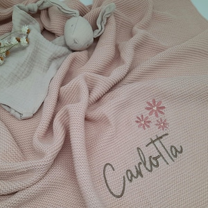 Puede incluir: Una manta de beb&eacute; de punto rosa claro con el nombre "Carlotta" bordado en cursiva, junto con tres flores bordadas. Tambi&eacute;n se ve un conejito de peluche y una muselina.