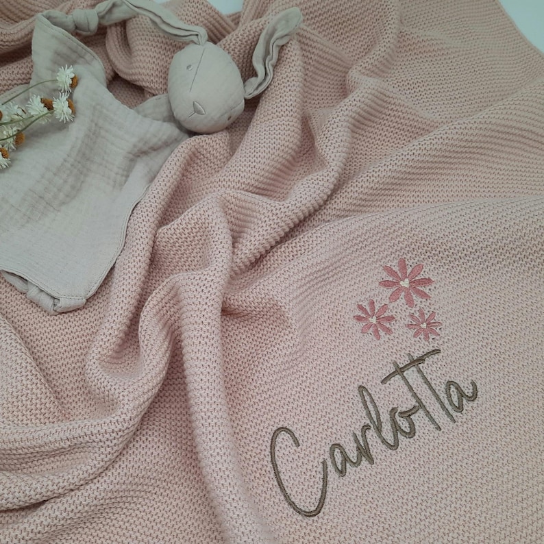 Puede incluir: Una manta de beb&eacute; de punto rosa claro con el nombre "Carlotta" bordado en cursiva, junto con tres flores bordadas. Tambi&eacute;n se ve un conejito de peluche y una muselina.