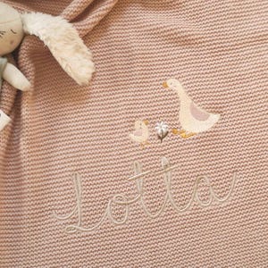 Puede incluir: Una manta de beb&eacute; de punto rosa con gansos bordados y el nombre "Lotta" en cursiva. Un juguete de peluche de conejito es parcialmente visible. La manta tiene una apariencia texturizada.