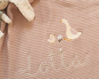 Puede incluir: Una manta de punto rosa empolvado con un bordado de un ganso, un pollito y una flor. El nombre Lotta está bordado en letra cursiva debajo de los animales. Un peluche con pelaje de color crema es visible en la esquina.