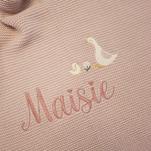 Puede incluir: Una manta de beb&eacute; de punto rosa claro con detalles bordados. La manta presenta un dise&ntilde;o de ganso y gansito con flores, y el nombre "Maisie" bordado en hilo rosa a juego. La manta tiene un aspecto texturizado.