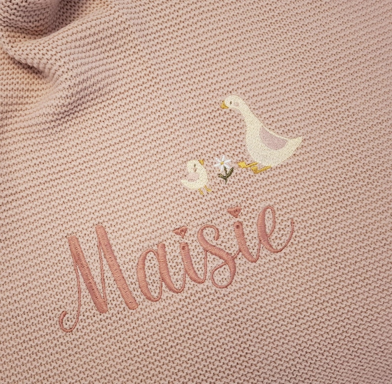 Puede incluir: Una manta de beb&eacute; de punto rosa claro con detalles bordados. La manta presenta un dise&ntilde;o de ganso y gansito con flores, y el nombre "Maisie" bordado en hilo rosa a juego. La manta tiene un aspecto texturizado.
