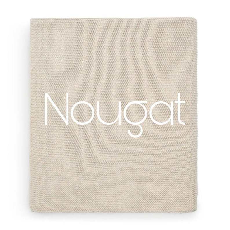 Puede incluir: Una manta de punto beige doblada con la palabra "Nougat" en escritura blanca en la parte delantera. La manta tiene un aspecto texturizado y est&aacute; hecha de un material suave. El color neutro la hace adecuada para varios estilos de decoraci&oacute;n del hogar.