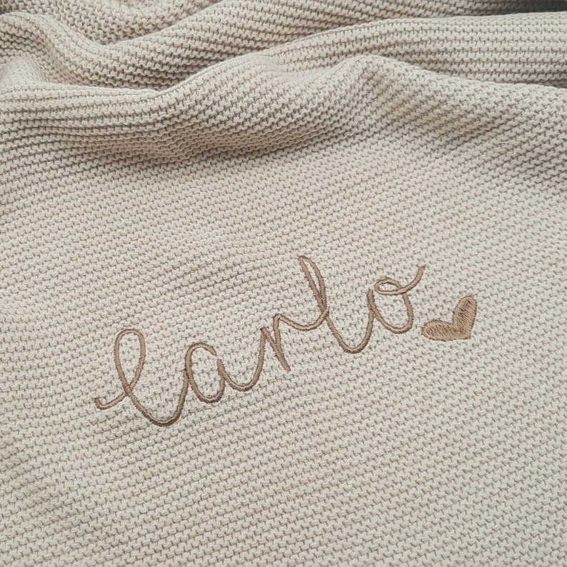 Puede incluir: Una manta beige tejida con el nombre "Carlo" bordado en cursiva, acompa&ntilde;ado de un peque&ntilde;o coraz&oacute;n. La manta tiene un aspecto texturizado y est&aacute; doblada, creando sombras suaves.