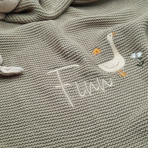 Op de afbeelding: Een saliegroene gebreide deken met een geborduurde witte gans, een kleine oranje bloem en een witte bloem. De naam Finn is in wit cursief schrift onder de gans geborduurd. Een beige knuffel met geknoopte ledematen is aan de deken bevestigd.