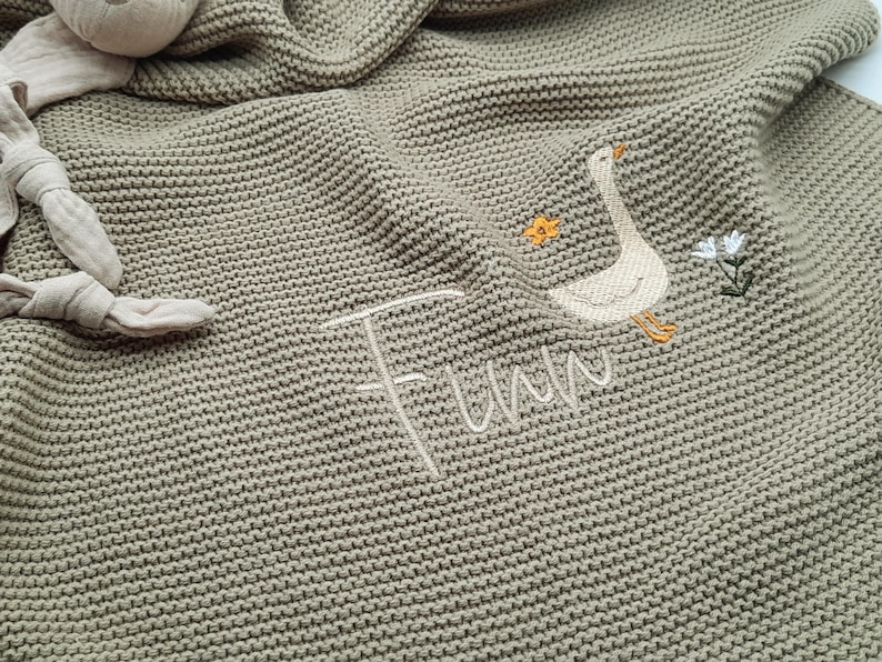 Op de afbeelding: Een saliegroene gebreide deken met een geborduurde witte gans, een kleine oranje bloem en een witte bloem. De naam Finn is in wit cursief schrift onder de gans geborduurd. Een beige knuffel met geknoopte ledematen is aan de deken bevestigd.