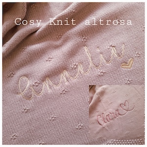Puede incluir: Una manta de punto suave, de color rosa empolvado, con un nombre bordado en cursiva. La manta tiene un patr&oacute;n texturizado con peque&ntilde;os puntos en relieve. Las palabras "Cosy Knit altrosa" est&aacute;n en la parte superior.