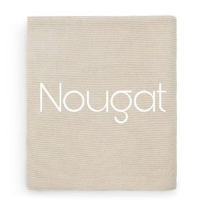 Op de afbeelding: Een rechthoekige, beige gebreide deken met een fijne, gelijkmatige textuur. Het woord "Nougat" is in grote, witte, schreefloze hoofdletters in het midden van het stoffen oppervlak gedrukt.