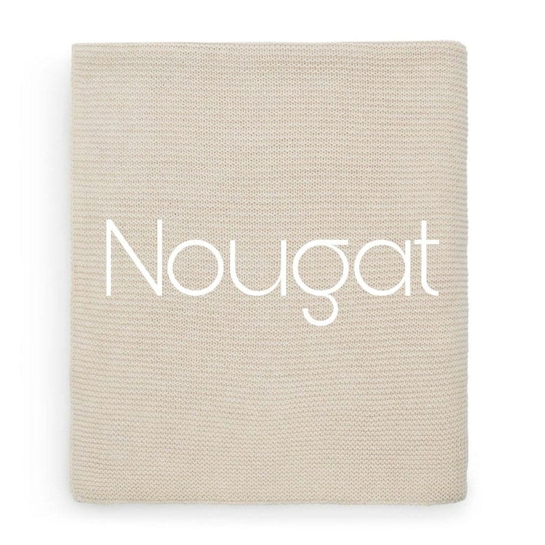 Op de afbeelding: Een rechthoekige, beige gebreide deken met een fijne, gelijkmatige textuur. Het woord "Nougat" is in grote, witte, schreefloze hoofdletters in het midden van het stoffen oppervlak gedrukt.