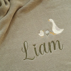 Puede incluir: Una manta de beb&eacute; de punto beige con el nombre "Liam" bordado en escritura verde. La manta presenta im&aacute;genes bordadas de un ganso, un pollito y una flor.
