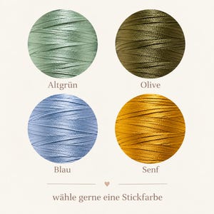Op de afbeelding: Vier ronde klossen borduurgaren in saliegroen, olijfgroen, lichtblauw en mosterdgeel. De tekstlabels bevatten Altgr&uuml;n, Olive, Blau, Senf en de zin w&auml;hle gerne eine Stickfarbe met een klein hartvormig icoontje in het midden.