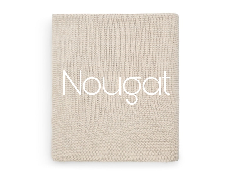 Puede incluir: Una manta tejida beige doblada con la palabra "Nougat" en escritura blanca en la parte delantera. La manta tiene un aspecto texturizado y probablemente est&aacute; hecha de un material suave.