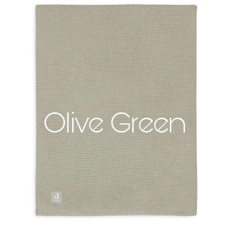 Op de afbeelding: Een rechthoekige gebreide deken in een gedempte olijfgroene tint met een fijne, gelijkmatige textuur. De tekst "Olive Green" is in het wit in het midden gedrukt en een klein vierkant label met de letter "J" is aan de onderhoek bevestigd.
