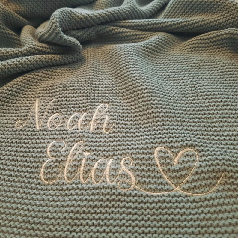 Puede incluir: Una manta de punto en color verde azulado apagado. La manta presenta los nombres bordados "Noah" y "Elias" en cursiva, junto con un s&iacute;mbolo de coraz&oacute;n. La manta tiene un aspecto texturizado.