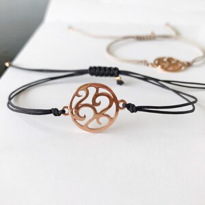 May include: A black cord bracelet with a rose gold circular charm featuring a decorative swirl design.