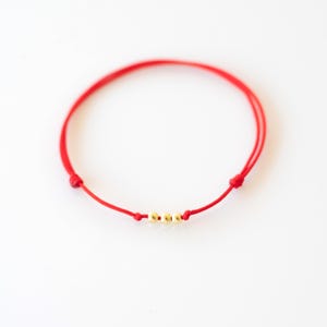 Könnte beinhalten: Ein rotes Armband mit drei kleinen goldenen Perlen in der Mitte. Das Armband ist an jedem Ende geknotet und vor einem weißen Hintergrund platziert. Schlichtes Design.