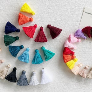 Op de afbeelding: Een verzameling kleurrijke kwastjes in diverse tinten, waaronder geel, oranje, rood, roze en blauw. Elk kwastje heeft een kleine, goudkleurige dop. De kwastjes zijn in een cirkelvorm gerangschikt, met enkele verspreid. Een kaart met tekst staat op de achtergrond.