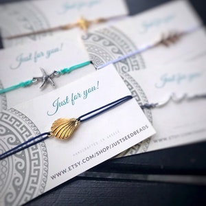 Może przedstawiać: Zestaw ręcznie robionych bransoletek z zawieszką w kształcie muszli i tekstem "Just for you!" na białej kartce. Bransoletki są wykonane z niebieskiego i turkusowego sznurka i mają złotą zawieszkę w kształcie muszli. Kartka zawiera również tekst "Handcrafted in Greece" i adres strony internetowej.