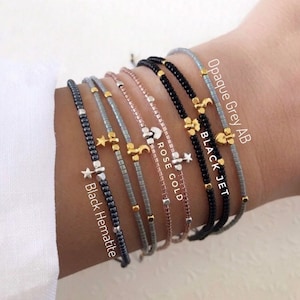 May include: A stack of seven beaded bracelets with different colored beads and charms. The charms include a star, crescent moon, and heart. The bracelets are labeled with the bead colors: Black Hematite, Opaque Grey AB, Black Jet, Rose Gold.