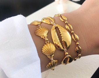 Puede incluir: Un conjunto de tres pulseras de conchas doradas. Las pulseras están hechas de pequeñas cuentas redondas y conchas más grandes. Los colgantes tienen forma de conchas y están conectados por un cordón beige delgado.