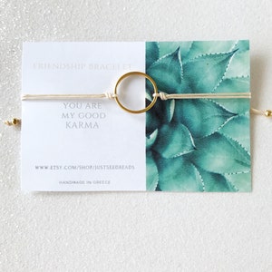 Peut inclure: Un bracelet d'amitié blanc avec un charme rond en or. Le bracelet porte le texte "You are my good karma" et "www.etsy.com/shop/justseedbeads" imprimé sur une carte blanche. La carte indique également "Handmade in Greece".