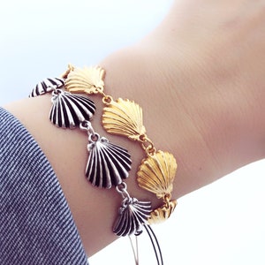 Pode incluir: Uma pulseira de concha dourada e prateada. A pulseira apresenta berloques de concha dourados e prateados alternados, ligados entre si. A pulseira é usada no pulso, com uma manga de jaqueta jeans ao fundo.