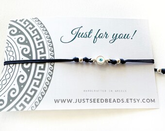 May include: A black string bracelet with a silver charm featuring a blue evil eye. The bracelet is on a white card with the text "Just for you!" and "www.justseedbeads.etsy.com".