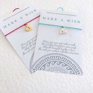 May include: Two "Make a Wish" bracelets on cards. One has a red string and a gold heart charm. The other has a teal string and a silver heart charm with the letter "A". The cards have text and a decorative border.