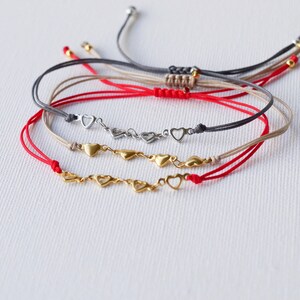 May include: Four friendship bracelets with heart charms. The bracelets are red, beige, and grey. The heart charms are gold and silver.