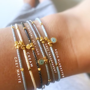 May include: A stack of six beaded bracelets with different colored beads and gold charms. The bracelets are labeled "Aqua Alabaster", "Grey Transparent", "Crystal Jet Lined", "Cream", and "Grey Matte".