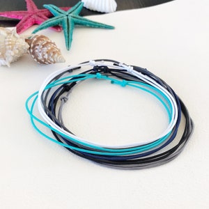 Könnte beinhalten: Ein Set aus acht bunten Armbändern in den Farben Weiß, Grau, Blau und Türkis. Die Armbänder bestehen aus dünnem, geflochtenem Band und sind zu einem Knoten gebunden.