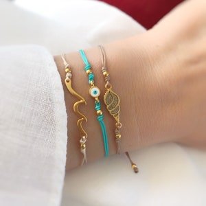 Può includere: Tre braccialetti dorati con ciondoli su un polso. I ciondoli sono un'onda, un occhio malvagio e una conchiglia. I braccialetti sono su cordini beige e turchese.
