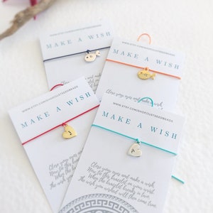 Op de afbeelding: Vier kaarten met de tekst "Make a Wish" erop. Elke kaart heeft een touwtje in een andere kleur met een bedel eraan. De bedels zijn een zilveren vis, een gouden vis, een zilveren hart en een gouden hart. De kaarten bevatten ook instructies over hoe je een wens kunt doen.