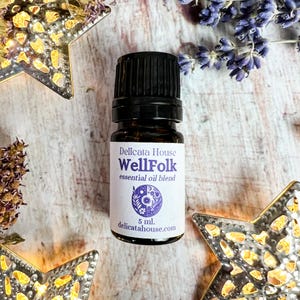May include: A small brown glass bottle with a black cap and a white label. The label says "Delicata House WellFolk essential oil blend" and has a purple flower design. The bottle is sitting on a wooden surface with silver stars and lavender flowers.