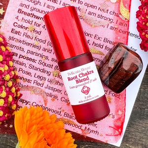 May include: A red glass roller bottle with a red label that reads "Delicata House Root Chakra Blend I am grounded & safe 5 ml. delicatahouse.com". The bottle is sitting on a pink and gold patterned surface with a red tiger's eye stone.