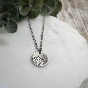May include: A silver necklace with a circular pendant depicting a mountain and water scene. The pendant hangs from a fine chain. The image is set against a white marble surface, with a green plant visible in the background.