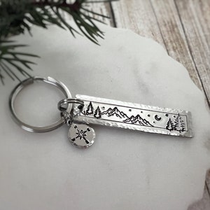 May include: Silver keychain with a compass charm and a rectangular tag featuring a mountain range, trees, a crescent moon, and stars.