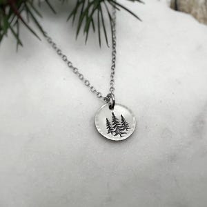 May include: A silver-toned necklace featuring a circular pendant with an etched design of three pine trees. The pendant hangs from a delicate chain. The necklace is displayed on a white, textured surface, with a sprig of greenery in the background.